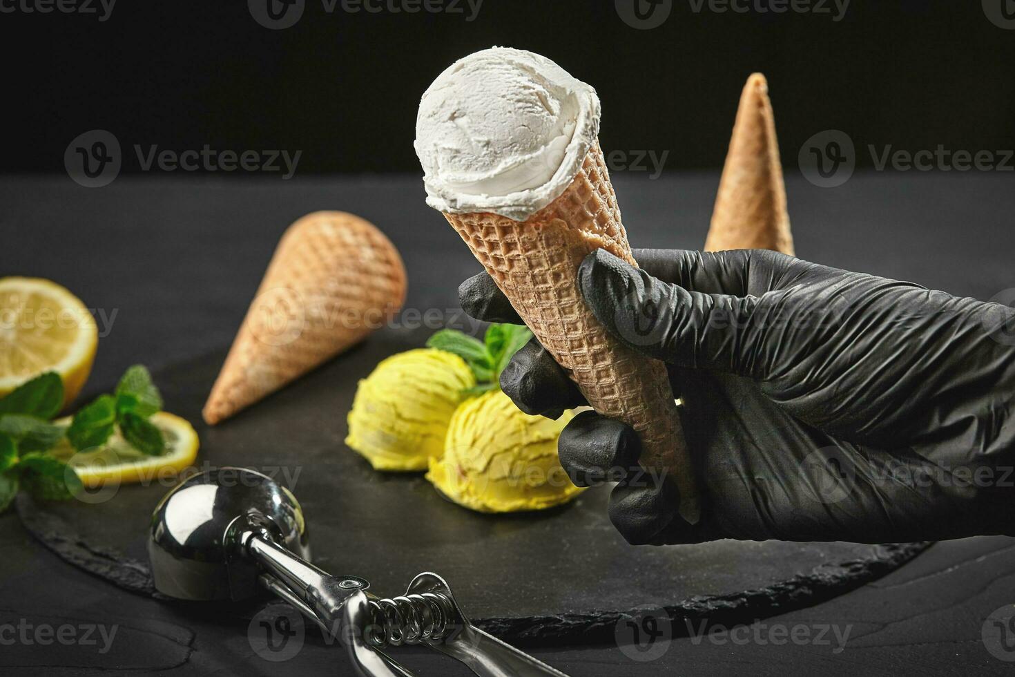 Hand in a dark glove is holding a waffle cone with a creamy ice cream on a background of a lemon sorbet decorated with mint. photo