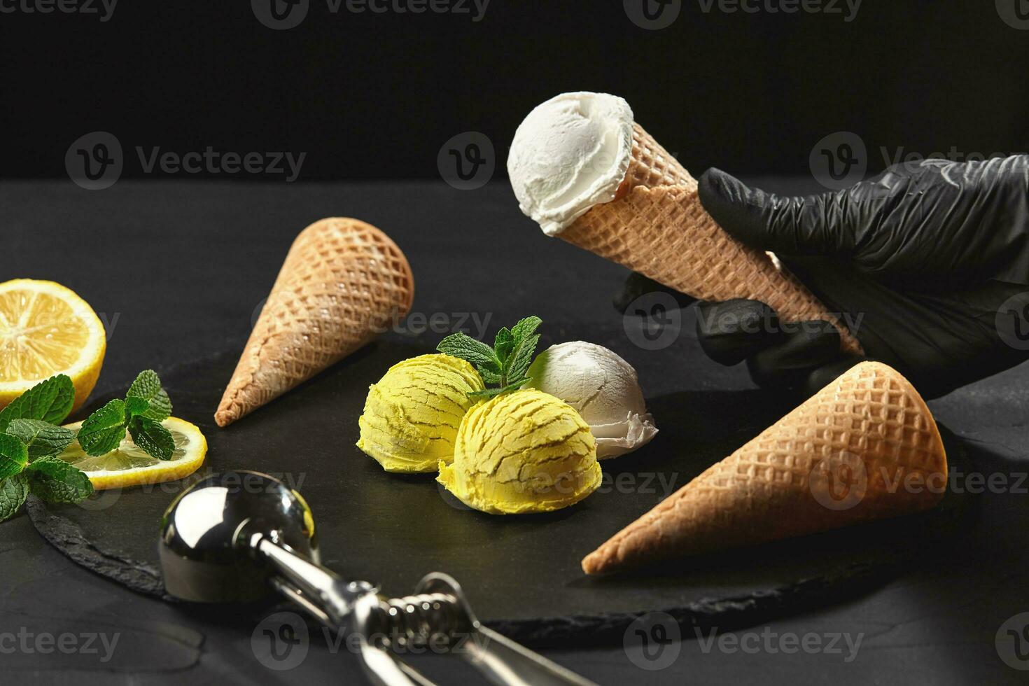 Hand in a dark glove is holding a waffle cone with a creamy ice cream on a background of a lemon sorbet decorated with mint. photo