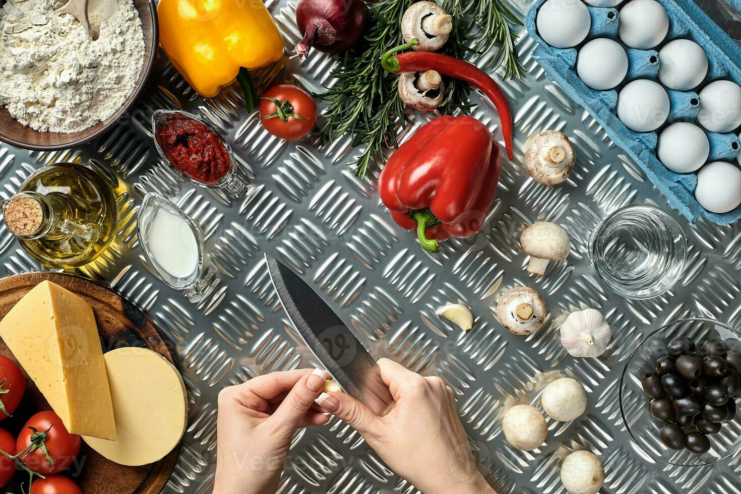 Female hand with knife on metal kitchen table, around lie ingredients, vegetables, and spices for pizza. photo