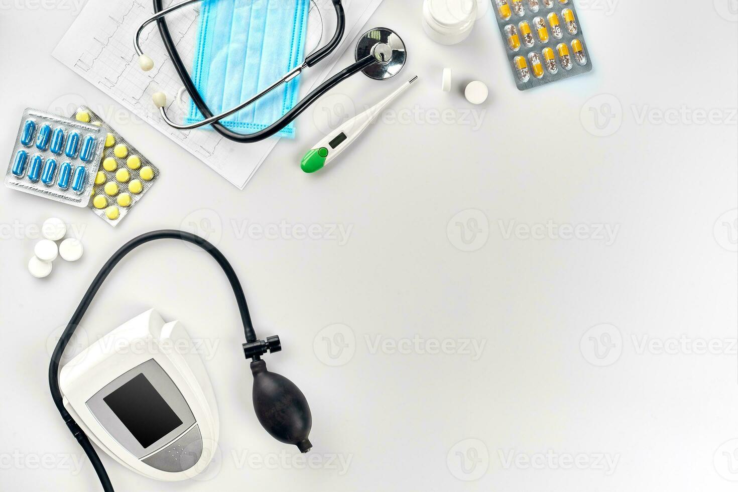 White electric tonometer with stethoscope, multicolored pills, mask and thermometer on white table. Top view photo