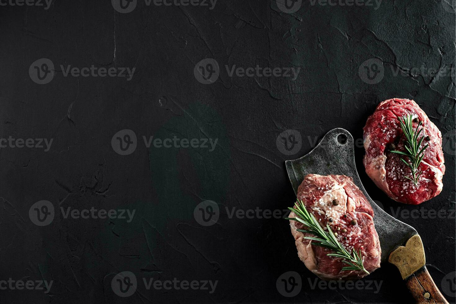 Two raw fresh ribeye steaks with salt, pepper, rosemary, garlic and an ax on black background photo