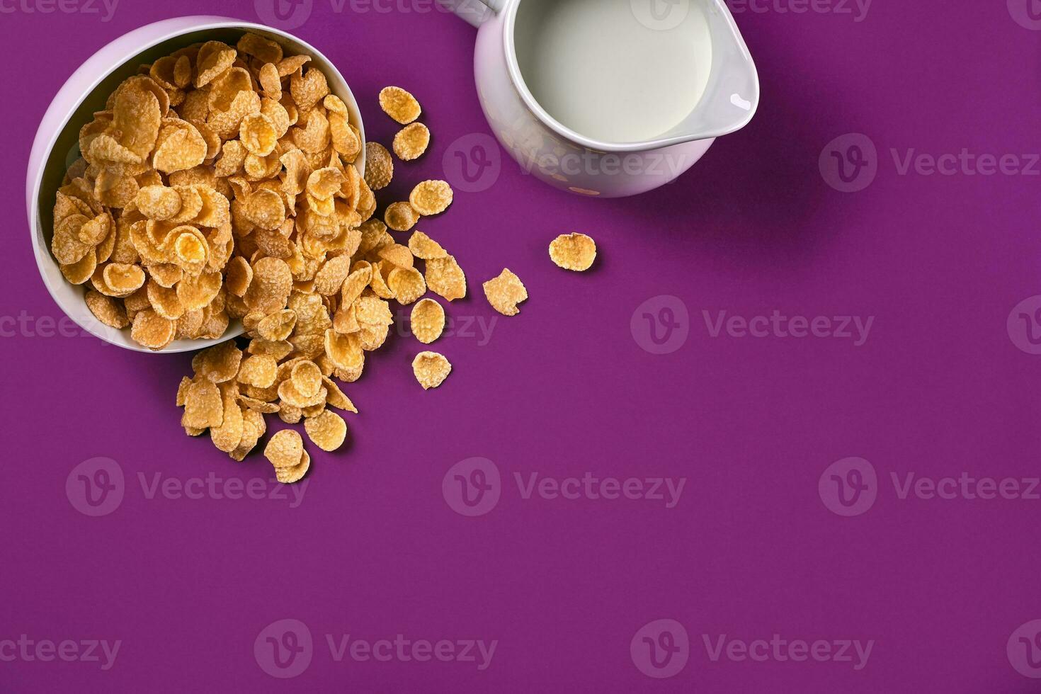 Bowl with corn flakes, jug of milk on purple background, top view photo