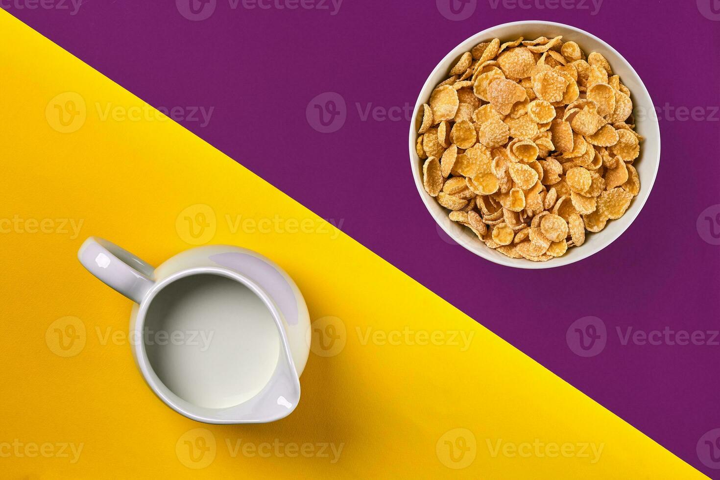 Bowl with corn flakes, jug of milk on purple and yellow background, top view photo