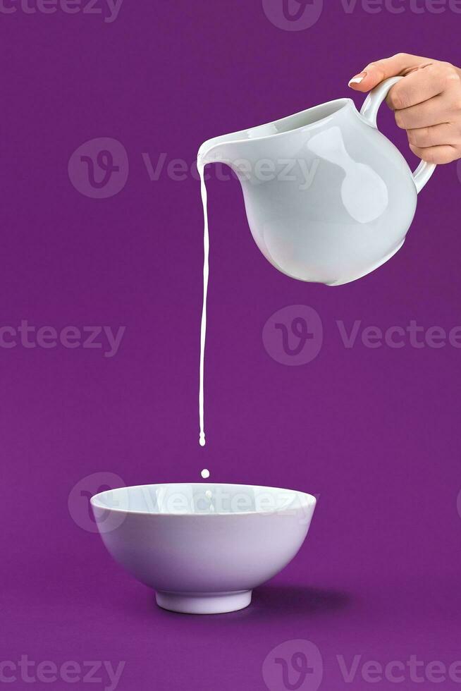 Partial view of hand pouring fresh organic milk from jug to bowl on purple background photo