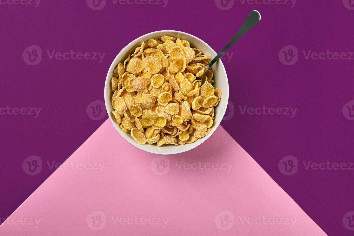 Bowl with corn flakes and spoon on purple and pink background, top view photo