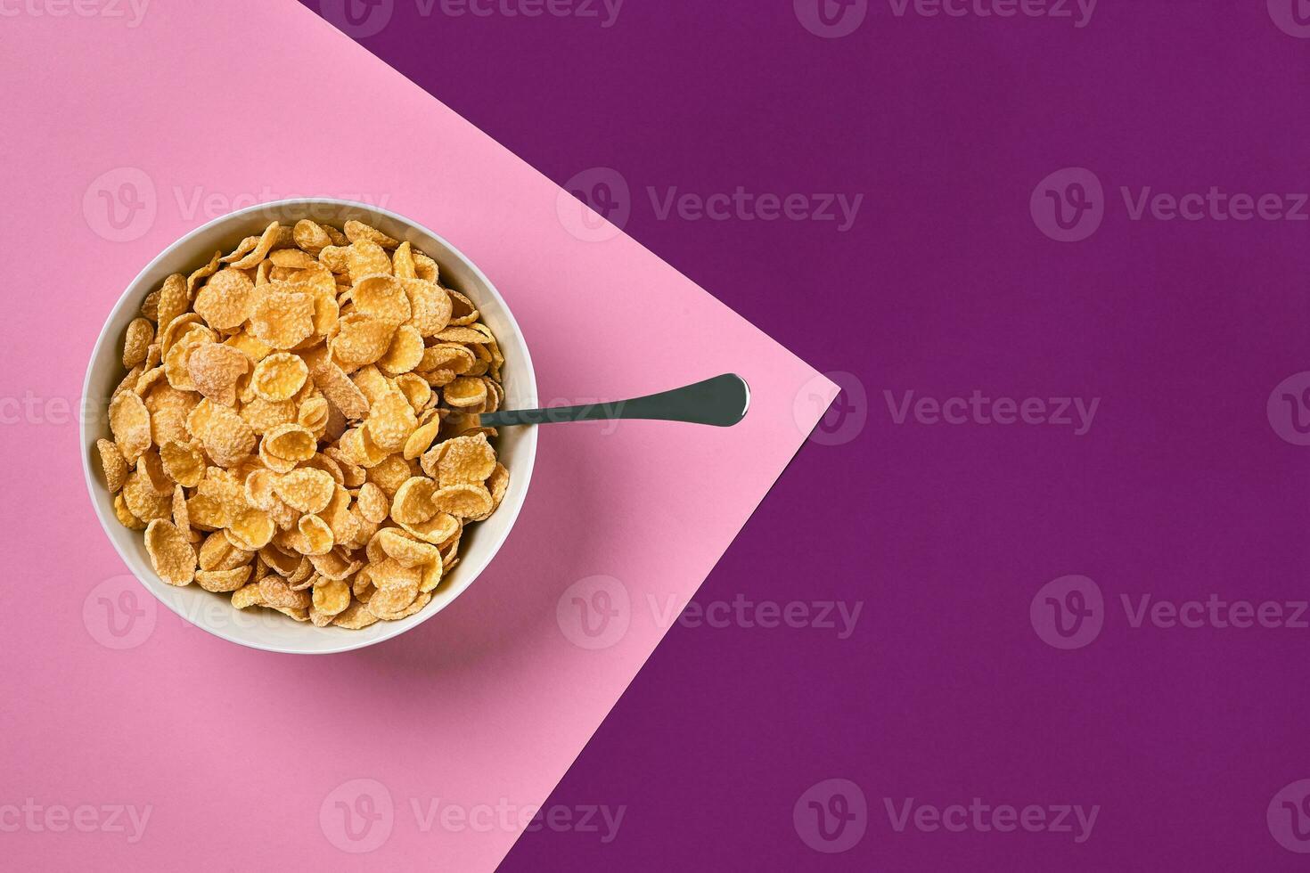 Bowl with corn flakes and spoon on purple and pink background, top view photo