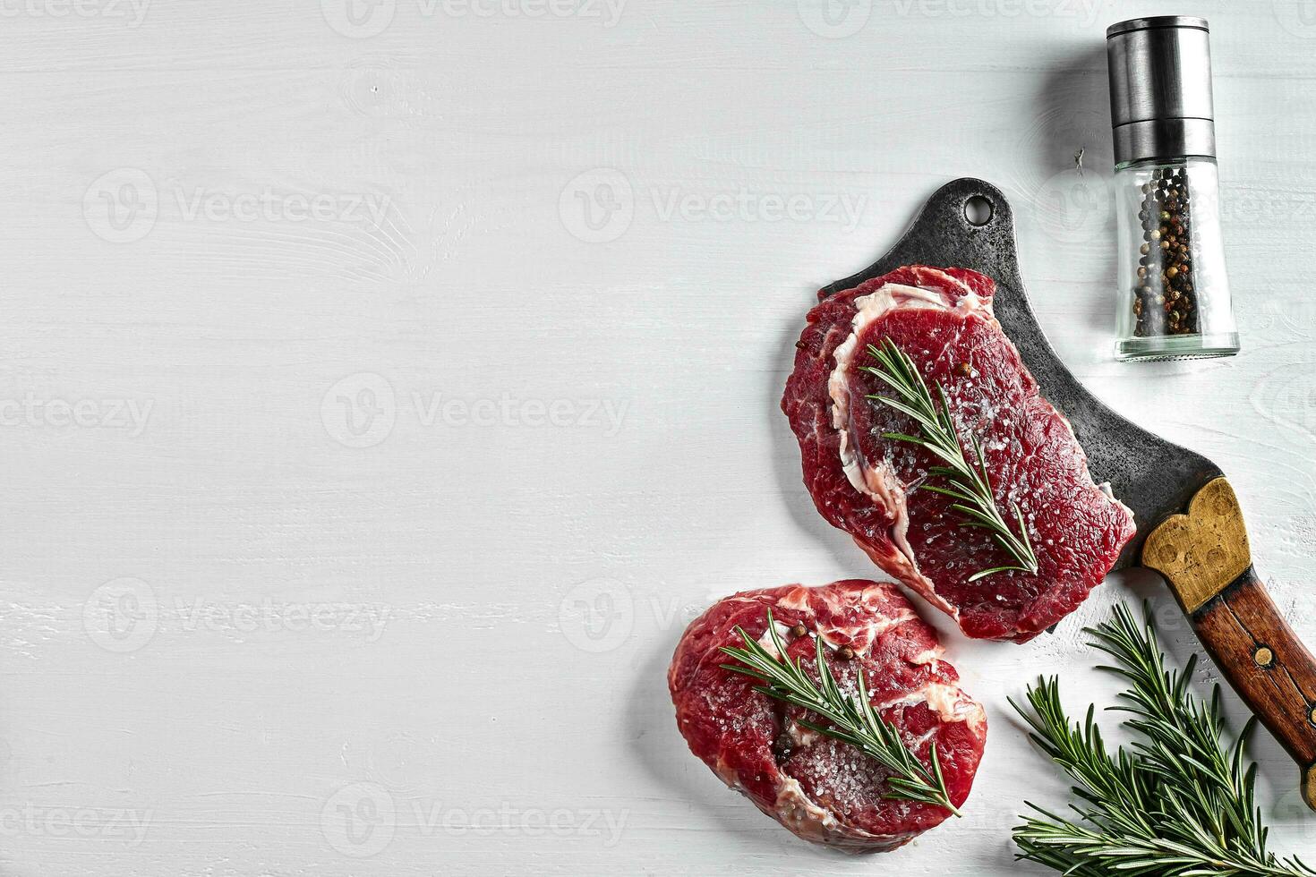 Two fresh raw steaks with herbs, garlic, olive oil, pepper, salt, rosemary and a kitchen ax on white background. Top view. photo