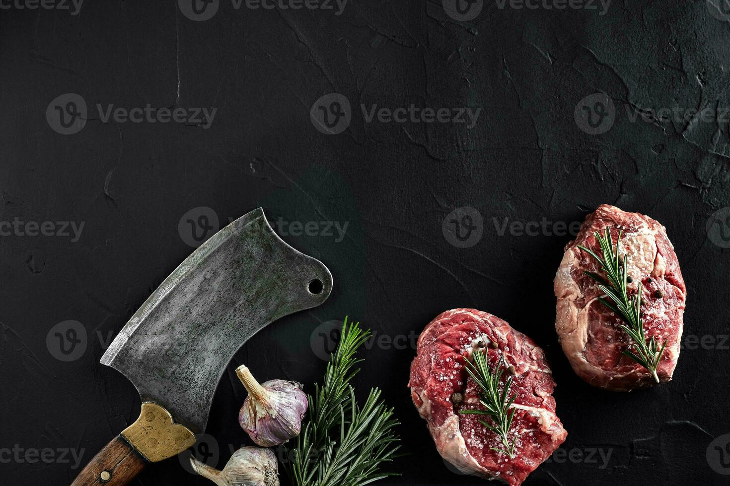 Two raw fresh ribeye steaks with salt, pepper, rosemary, garlic and an ax on black background photo