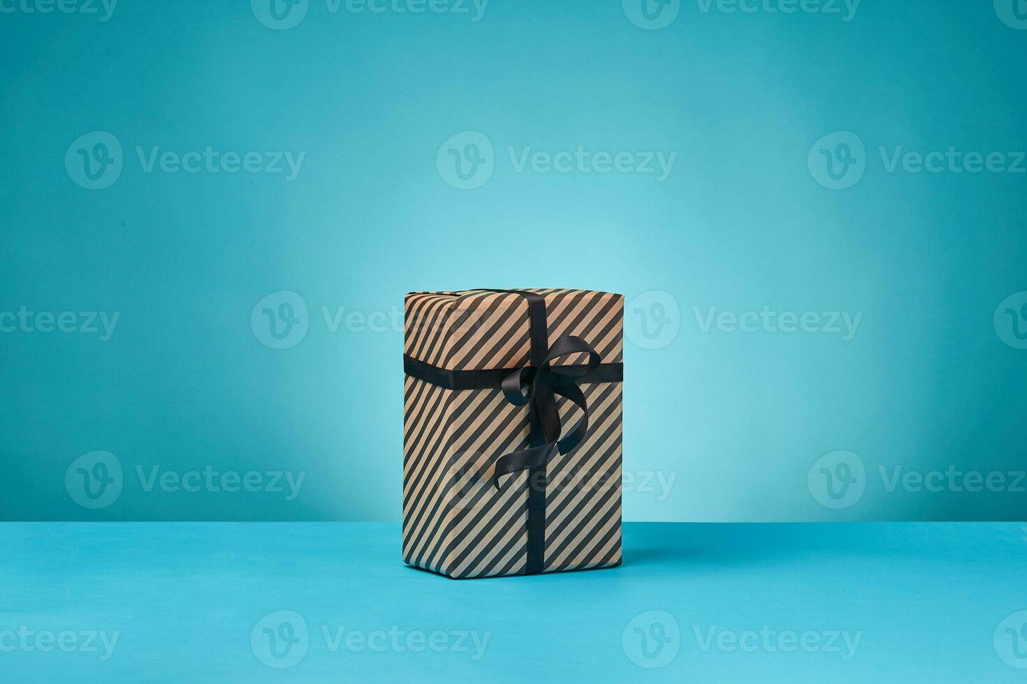 Striped brown paper present box tied with black ribbon and bow on a blue surface and background. Close-up, copy space. photo