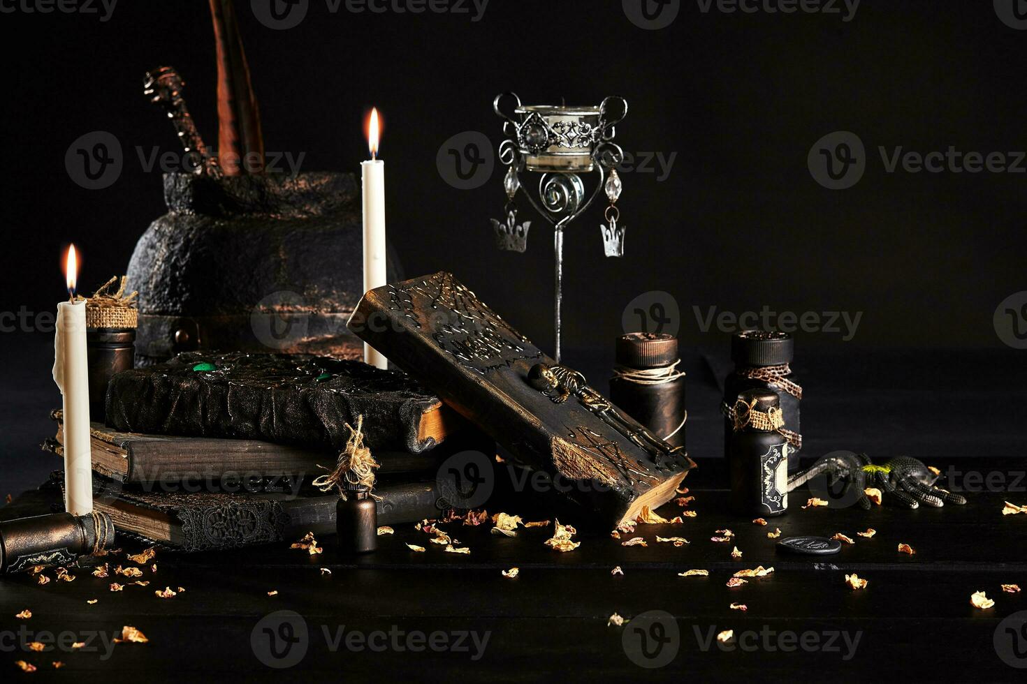 Books with spells, old pot, jars of potion are on a wooden dark table. Petals of dried roses are scattered nearby. Black background. Close-up shot. photo