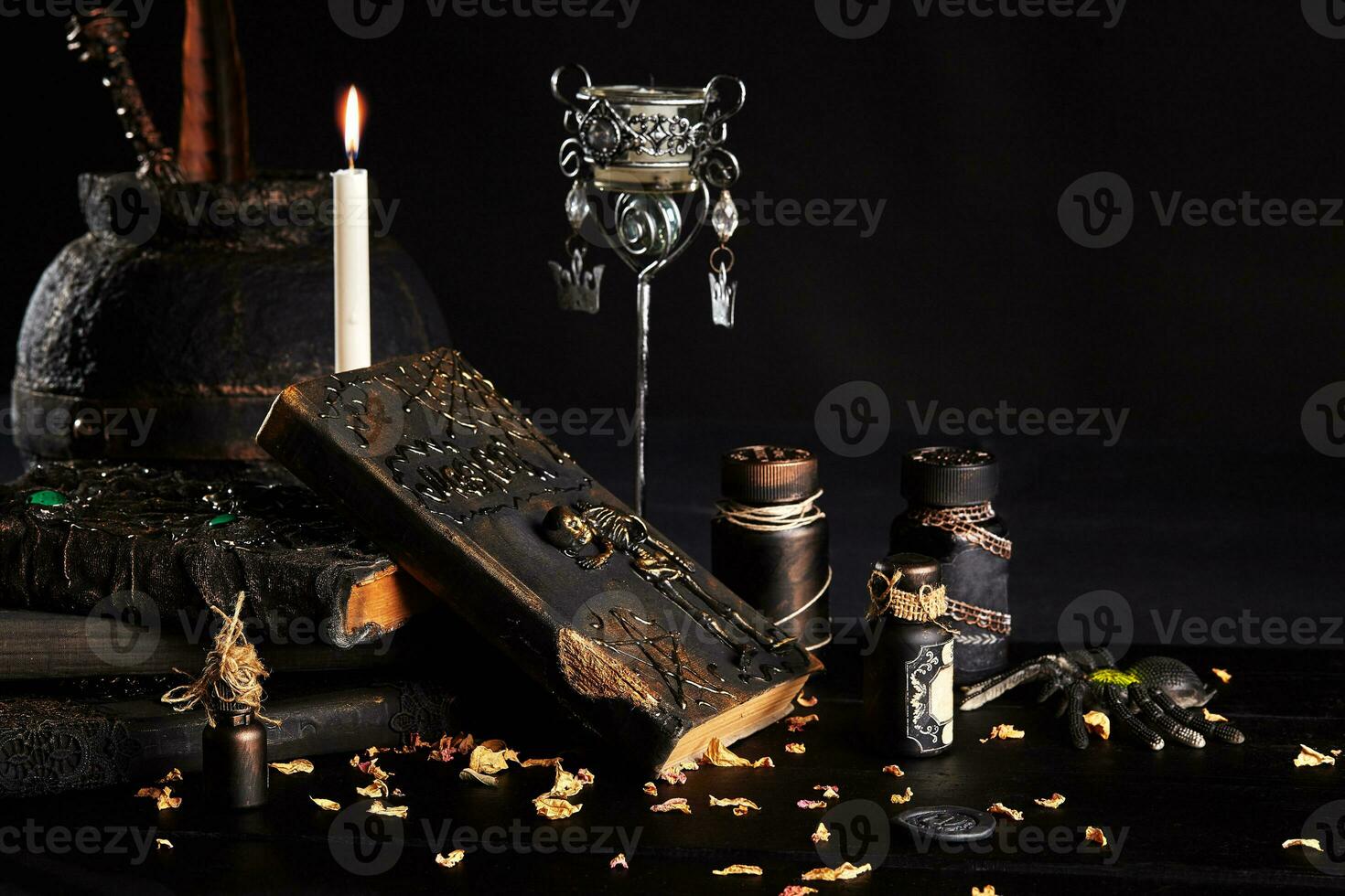 Books with spells, old pot, jars of potion are on a wooden dark table. Petals of dried roses are scattered nearby. Black background. Close-up shot. photo
