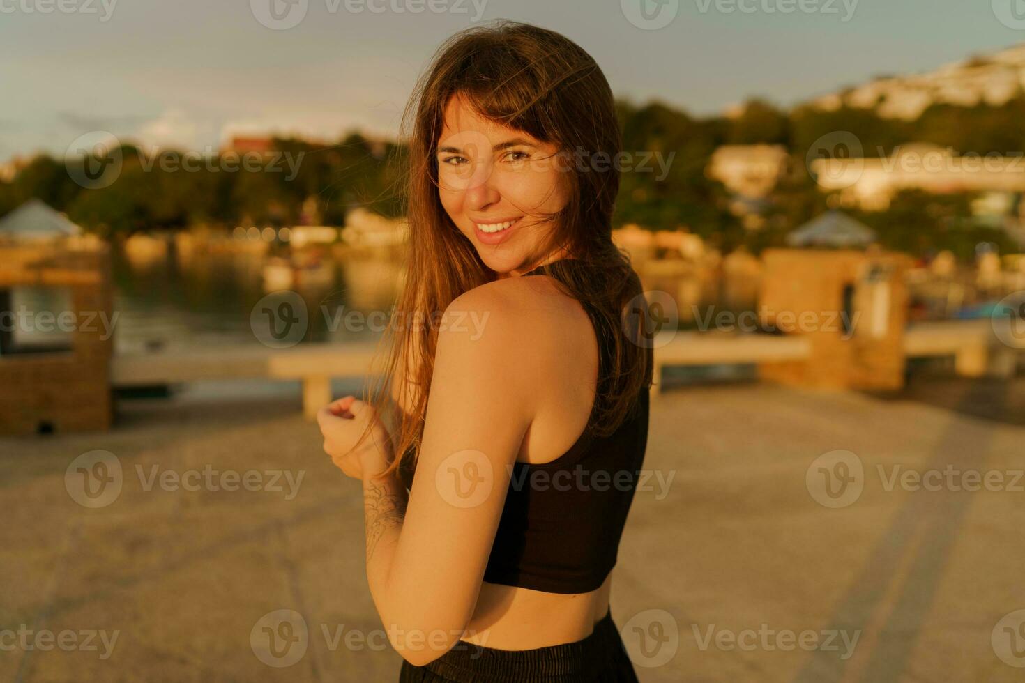 Beautiful brunette woman with stylish hairstyle walking outdoor   on the pier. Warm  sunser colors.  Travel and vacation concept. photo