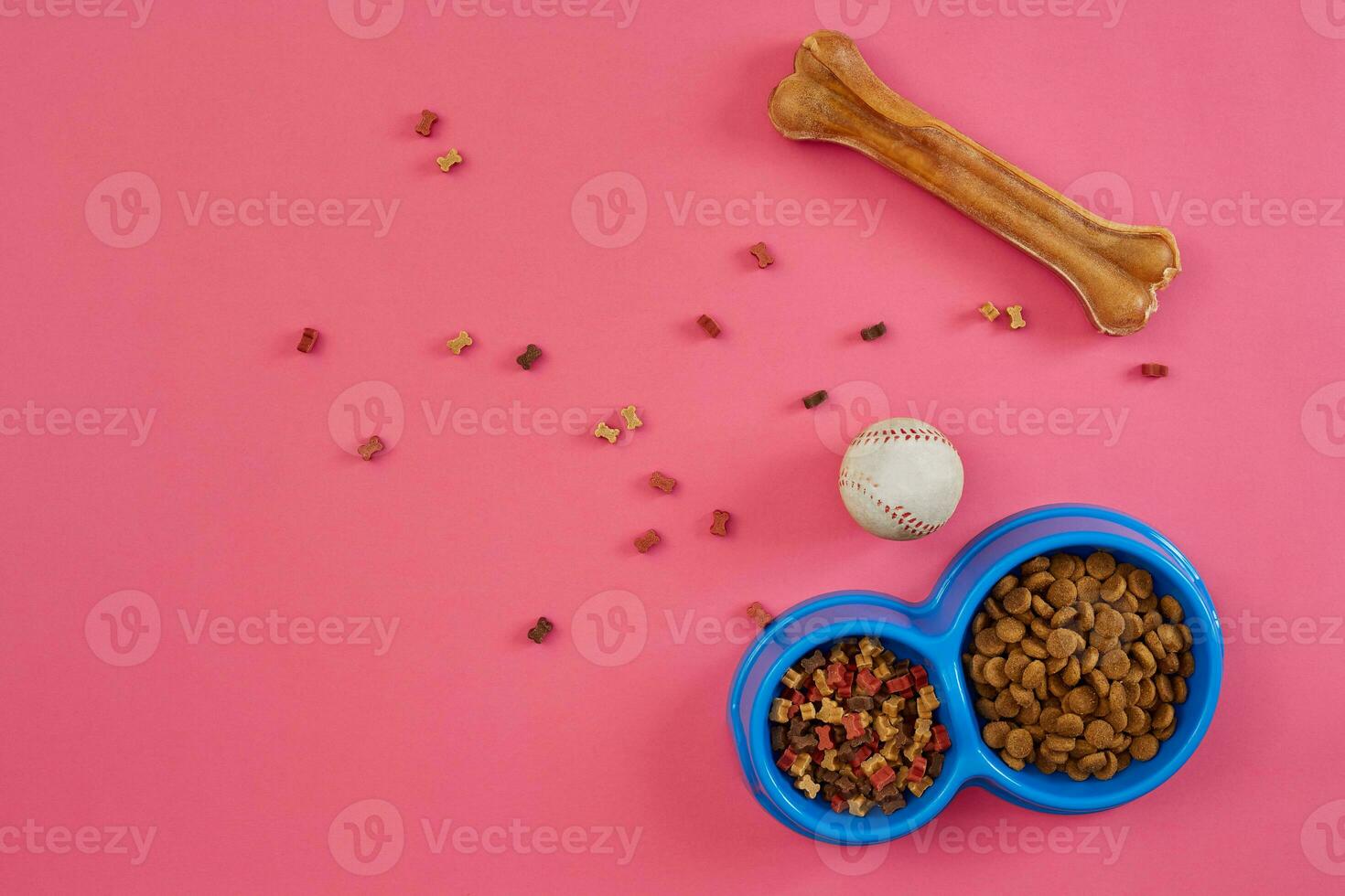 Dry pet food in bowl with a ball and dog bone on pink background top view photo