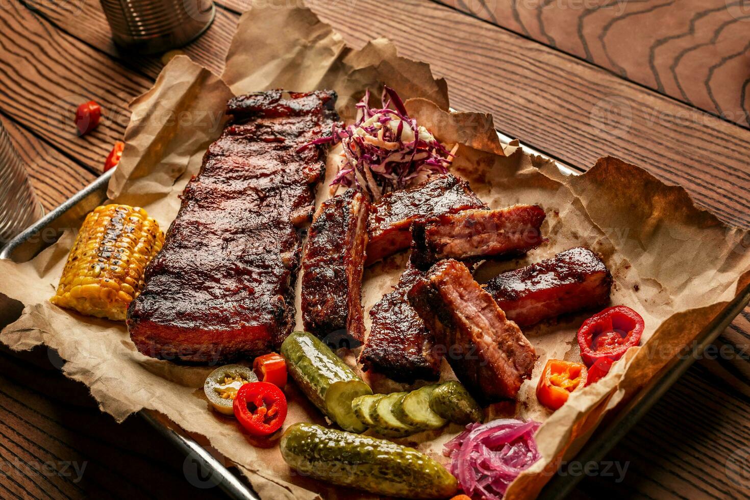 Grilled pork ribs served with grilled corn, salat, bbq sauce, salt pepper and cucumber on parchment paper on a wooden table. Top View photo