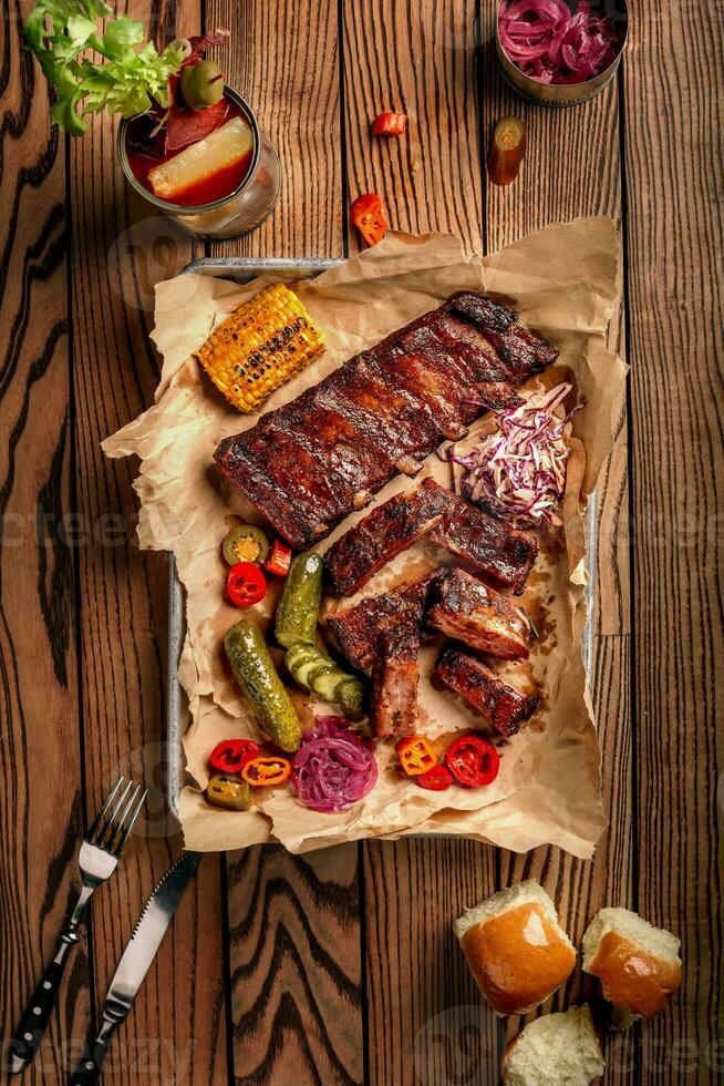 Grilled pork ribs served with grilled corn, salat, bbq sauce, salt pepper and cucumber on parchment paper on a wooden table. Top View photo