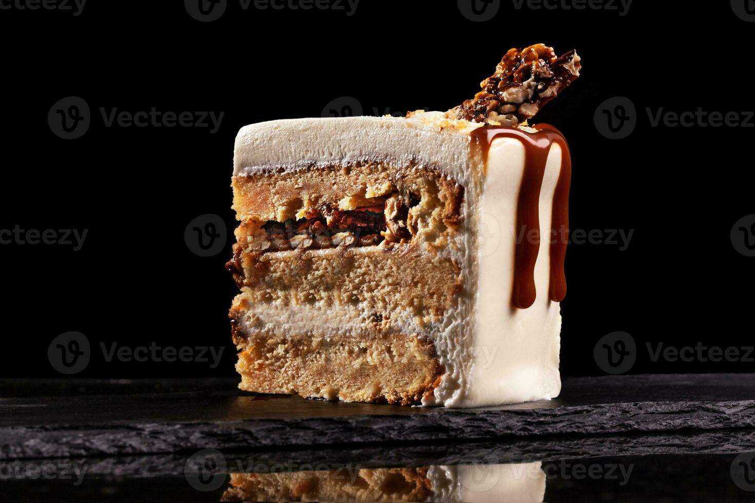 Piece of white cake with caramel and chocolate puffed rice on a black tray on black background photo