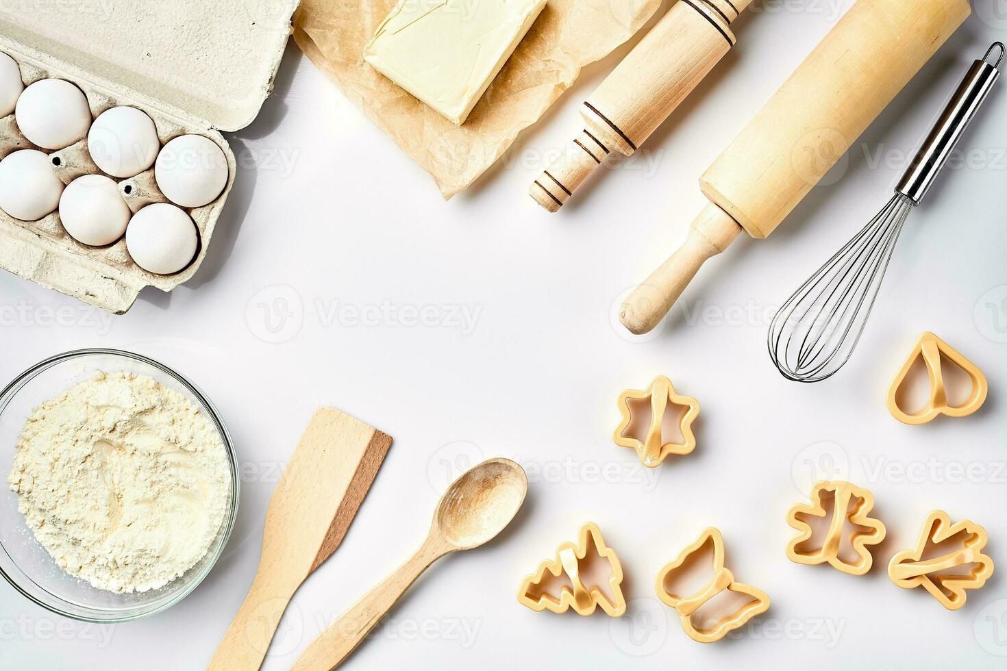 Bowl with wheat flour, rolling pin, whisk, eggs, butter, cookie cutters. Top view on a white table with a copy space photo