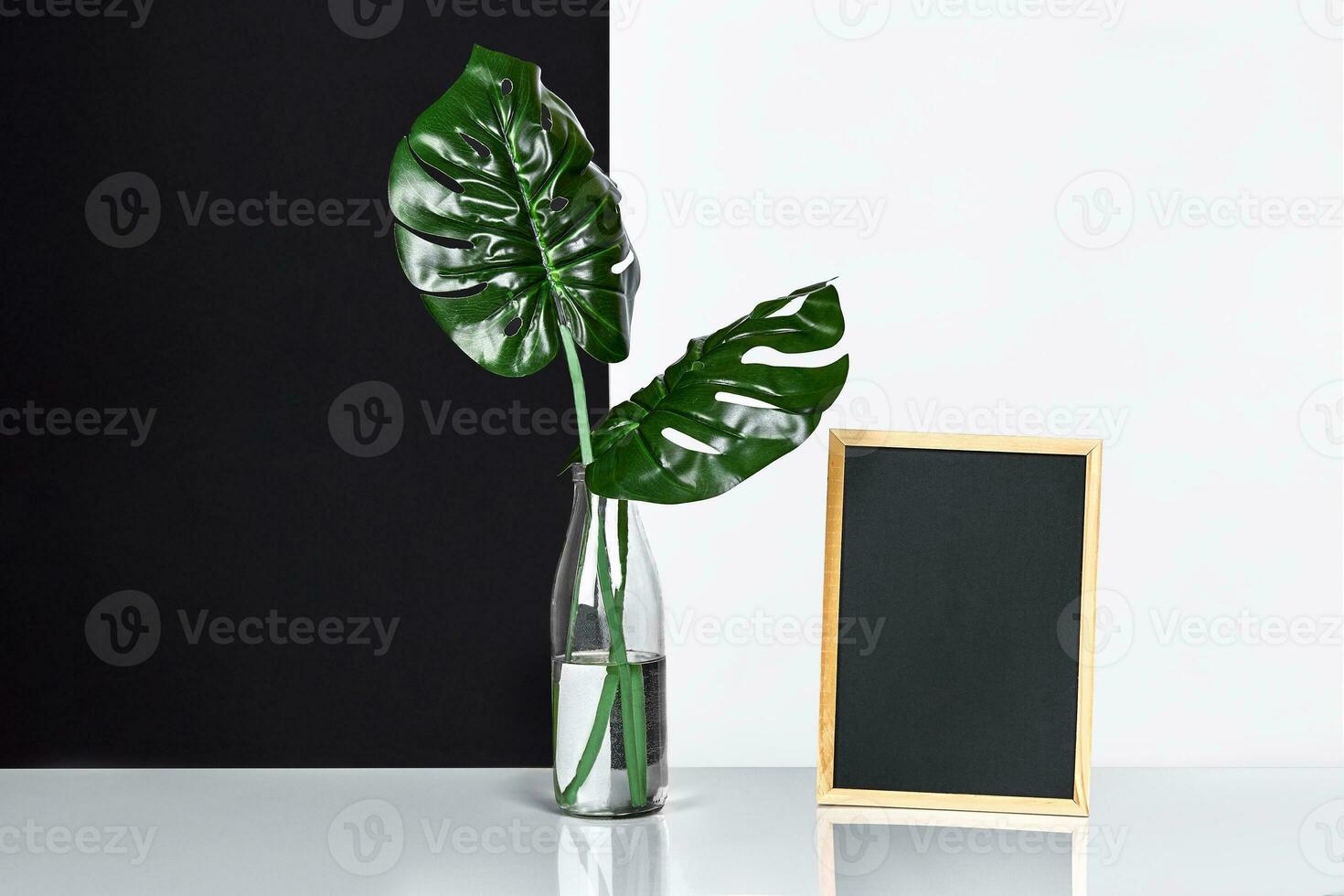 The stylish interior with mock up poster frame, leaves in glass bottle on table with black and white wall on background. photo