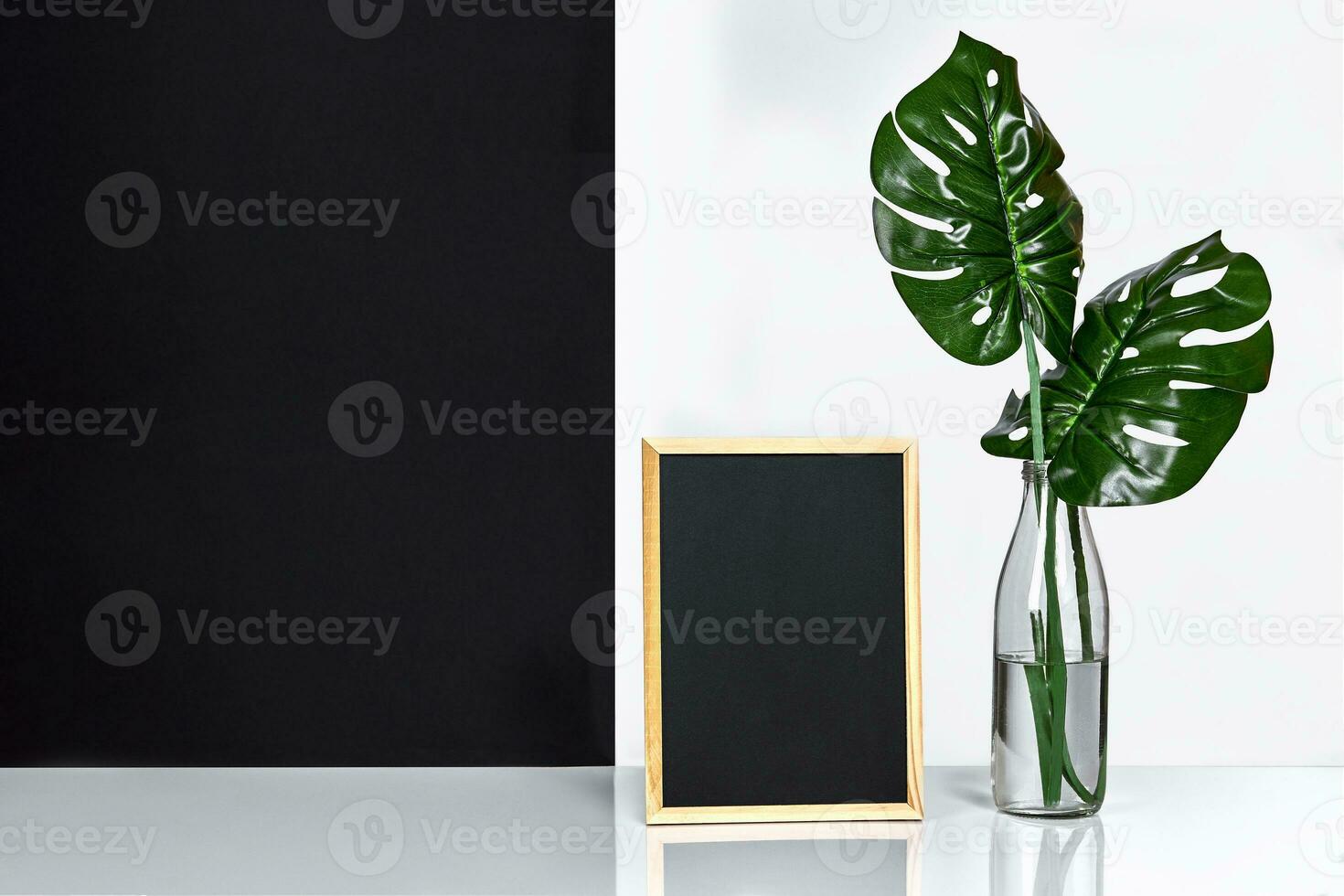 The stylish interior with mock up poster frame, leaves in glass bottle on table with black and white wall on background. photo