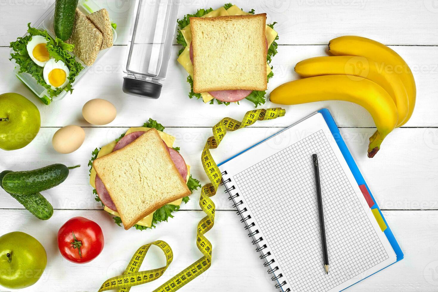 Top view planning notebook with copy space and healthy breakfast. Sandwich with ham and cheese, eggs, cucumber, tomato and bottle of water on white wooden table. photo