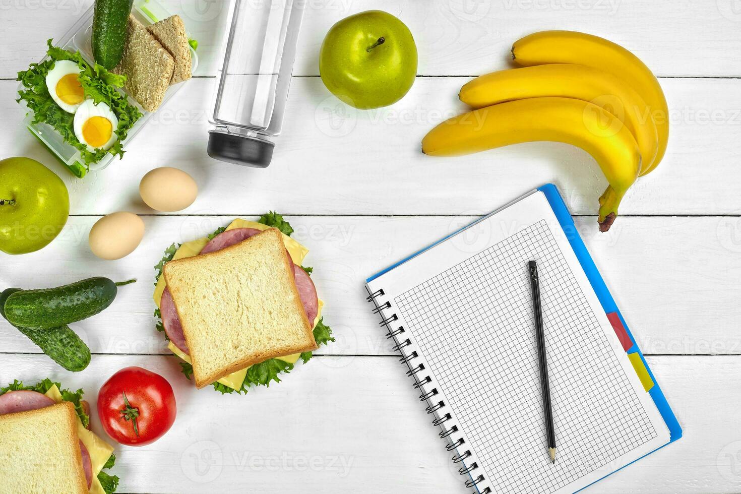 Top view planning notebook with copy space and healthy breakfast. Sandwich with ham and cheese, eggs, cucumber, tomato and bottle of water on white wooden table. photo