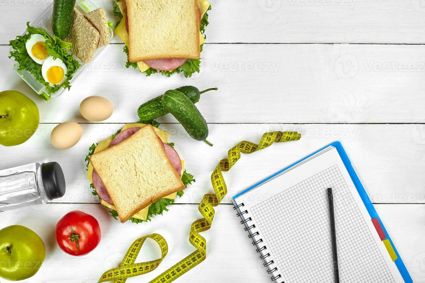Top view planning notebook with copy space and healthy breakfast. Sandwich with ham and cheese, eggs, cucumber, tomato and bottle of water on white wooden table. photo