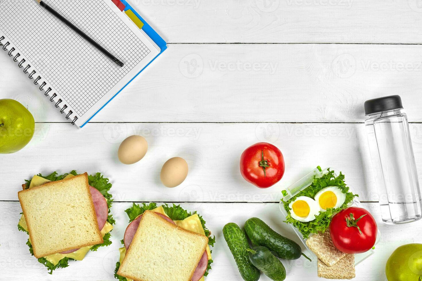 Top view planning notebook with copy space and healthy breakfast. Sandwich with ham and cheese, eggs, cucumber, tomato and bottle of water on white wooden table. photo
