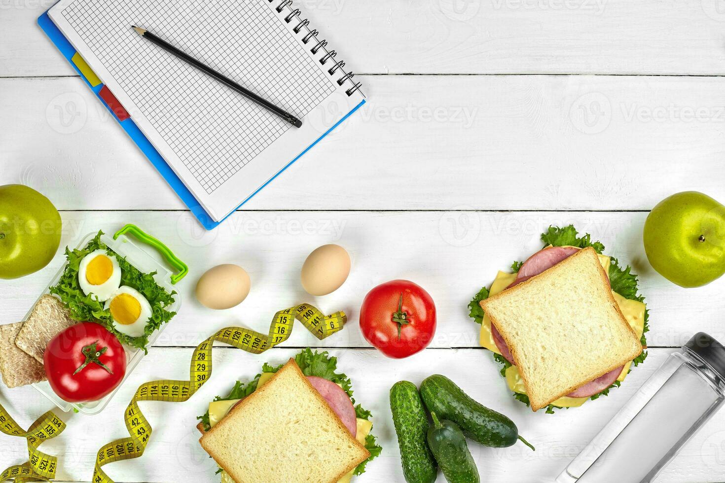 Top view planning notebook with copy space and healthy breakfast. Sandwich with ham and cheese, eggs, cucumber, tomato and bottle of water on white wooden table. photo