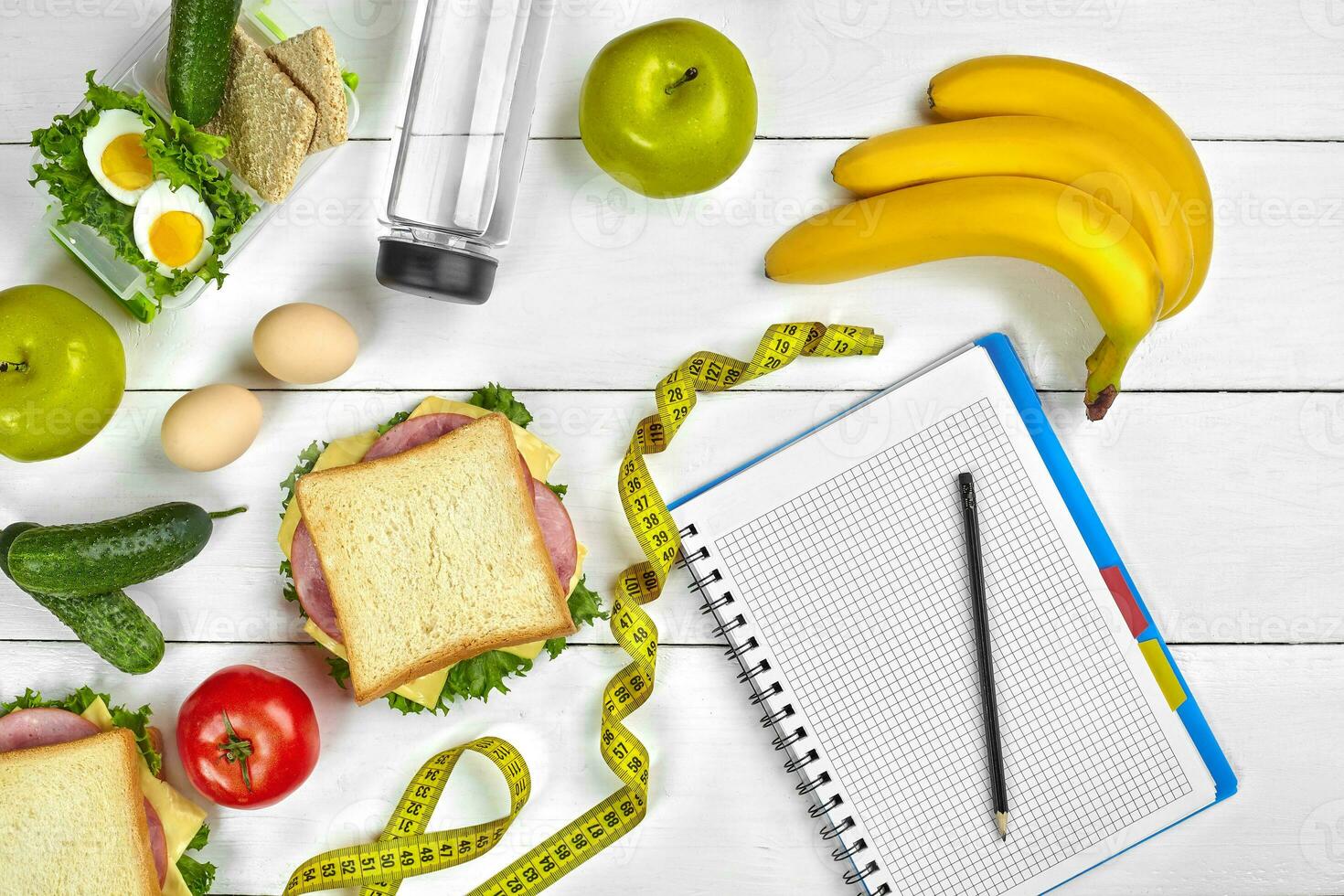Top view planning notebook with copy space and healthy breakfast. Sandwich with ham and cheese, eggs, cucumber, tomato and bottle of water on white wooden table. photo
