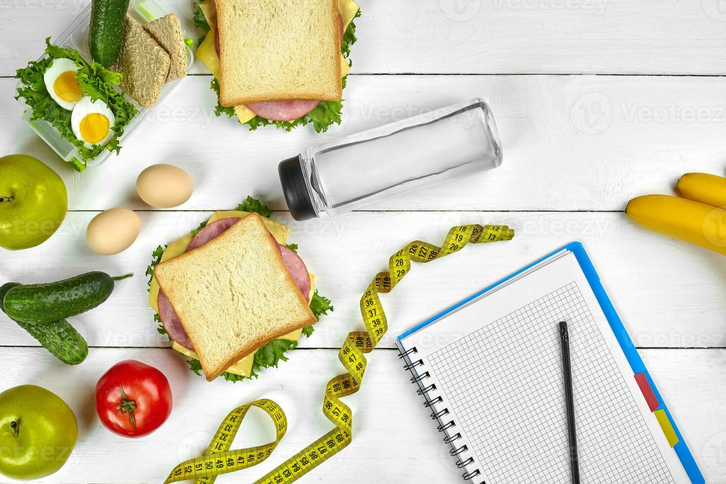 Top view planning notebook with copy space and healthy breakfast. Sandwich with ham and cheese, eggs, cucumber, tomato and bottle of water on white wooden table. photo