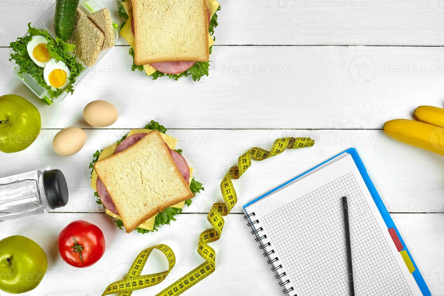 Top view planning notebook with copy space and healthy breakfast. Sandwich with ham and cheese, eggs, cucumber, tomato and bottle of water on white wooden table. photo