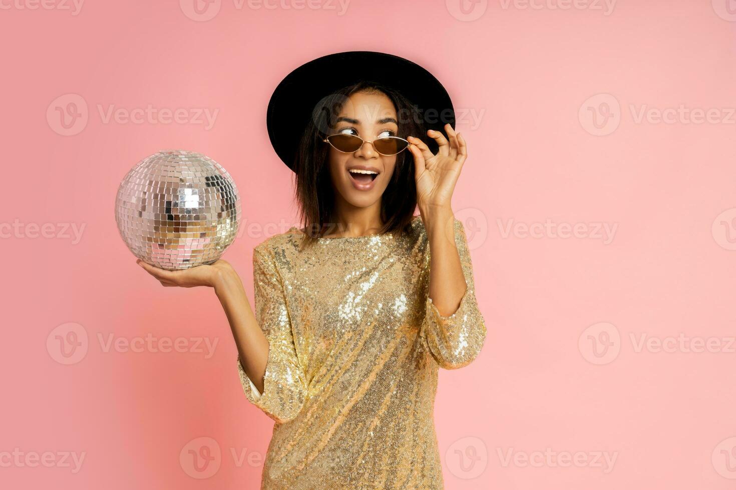 Celebrating woman in golden sequins dress  posing with disco baloon over pink bacground in studio. photo