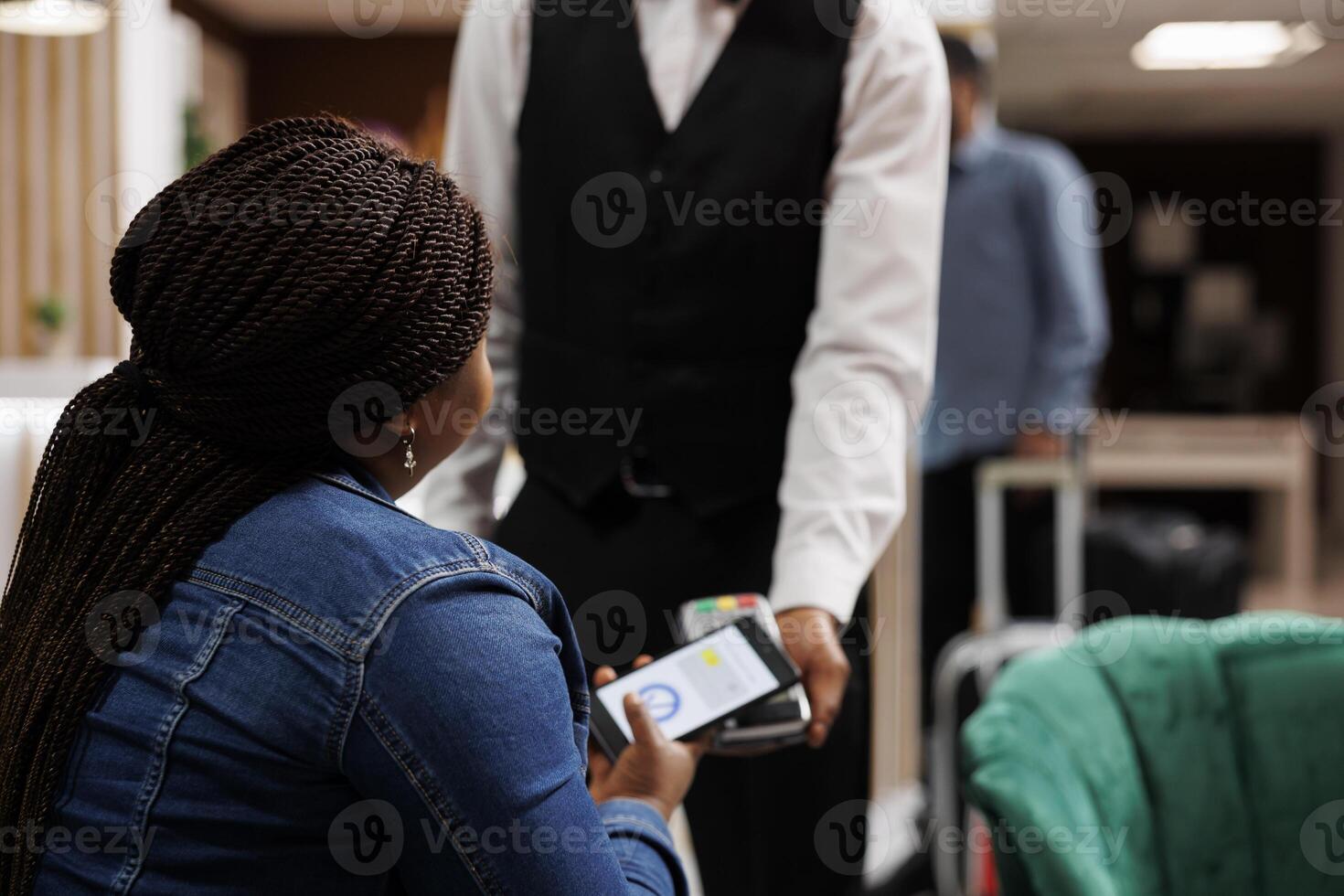 Female hotel guest making cashless payment using smartphone, close up. Tourist sitting in lobby paying for services with phone, waiter accepting contactless NFC payment from customer photo