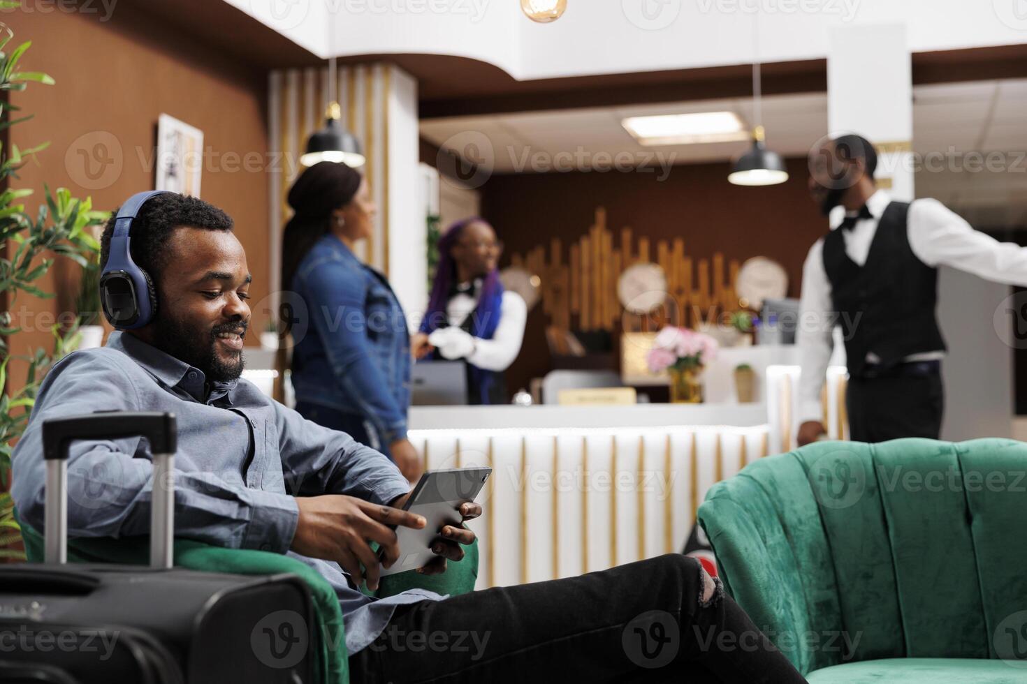 Happy African American guy hotel guest holding digital device making video call while relaxing in lobby. Relaxed black millennial man tourist wearing headphones using tablet waiting for checkin photo