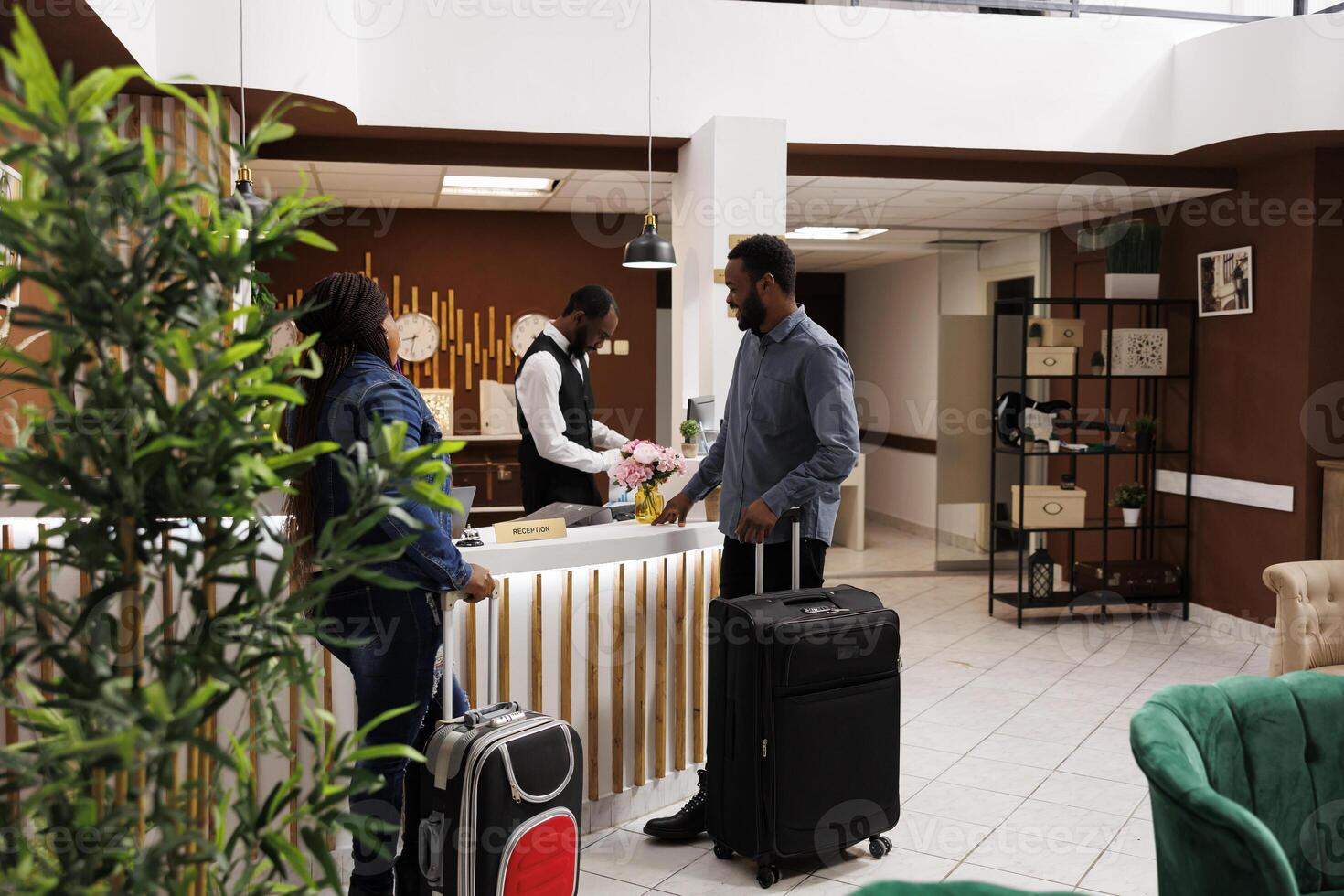 Happy black couple standing at hotel front desk with luggage, checking in at honeymoon resort, people waiting at reception for checkin. Receptionist helping guests with check out photo