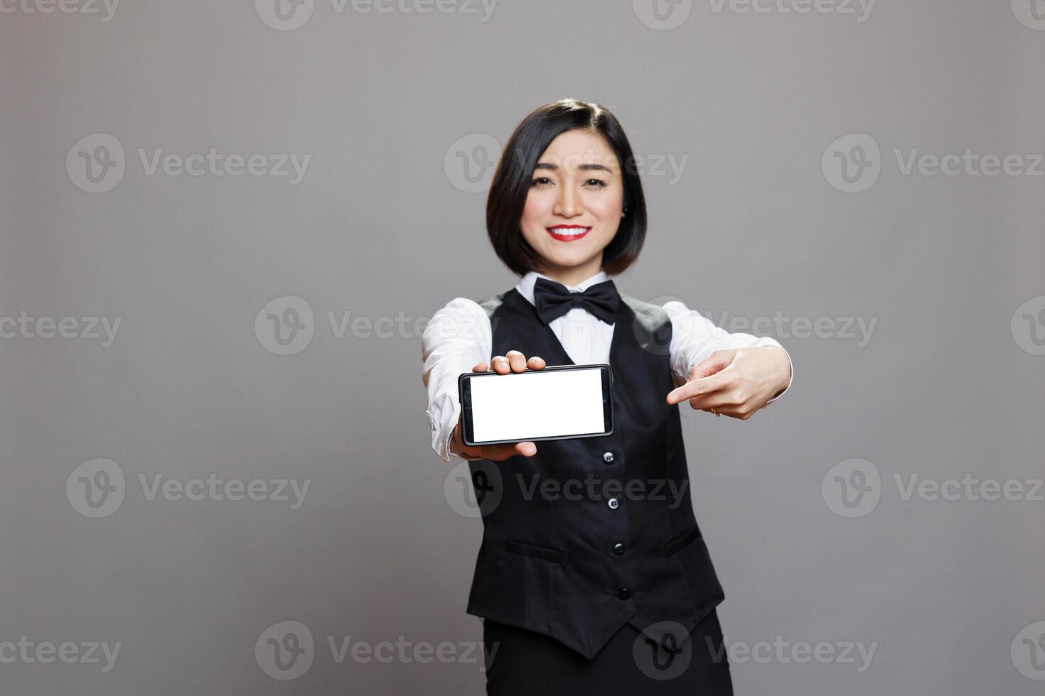 Cheerful asian receptionist showing smartphone blank screen, pointing with finger and looking at camera. Happy receptionist presenting mobile phone empty touchscreen portrait photo