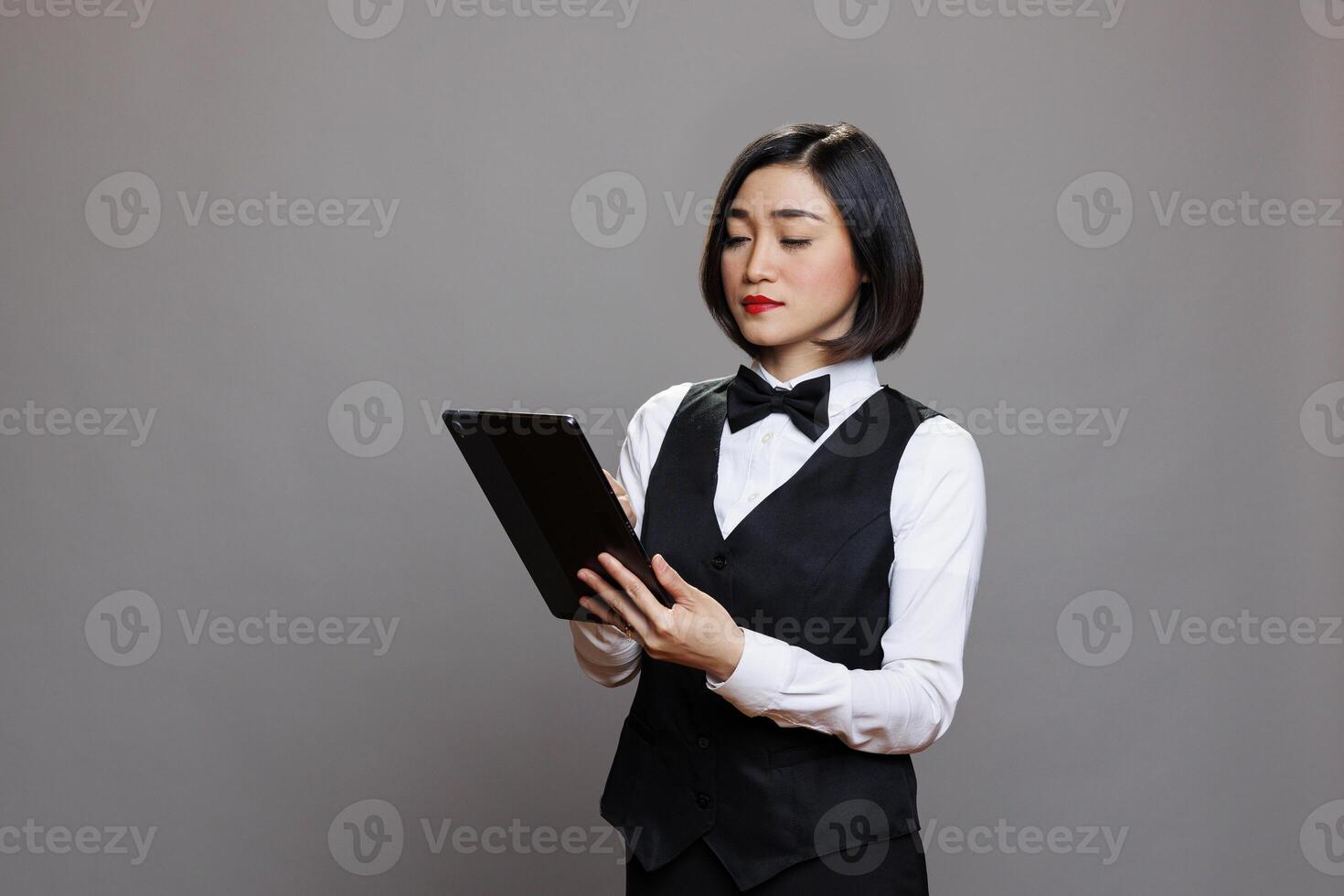 Confident asian waitress using digital tablet and checking orders online while posing in studio. Young woman restaurant receptionist wearing professional uniform holding portable gadget photo