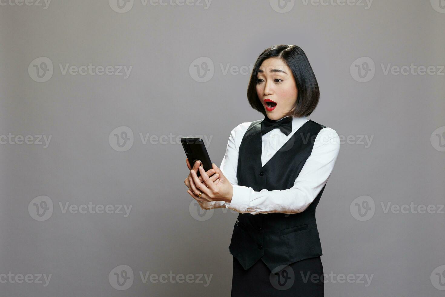Shocked happy receptionist checking customer satisfaction message on smartphone. Excited young asian waitress in black and white uniform reading client positive review on mobile phone photo
