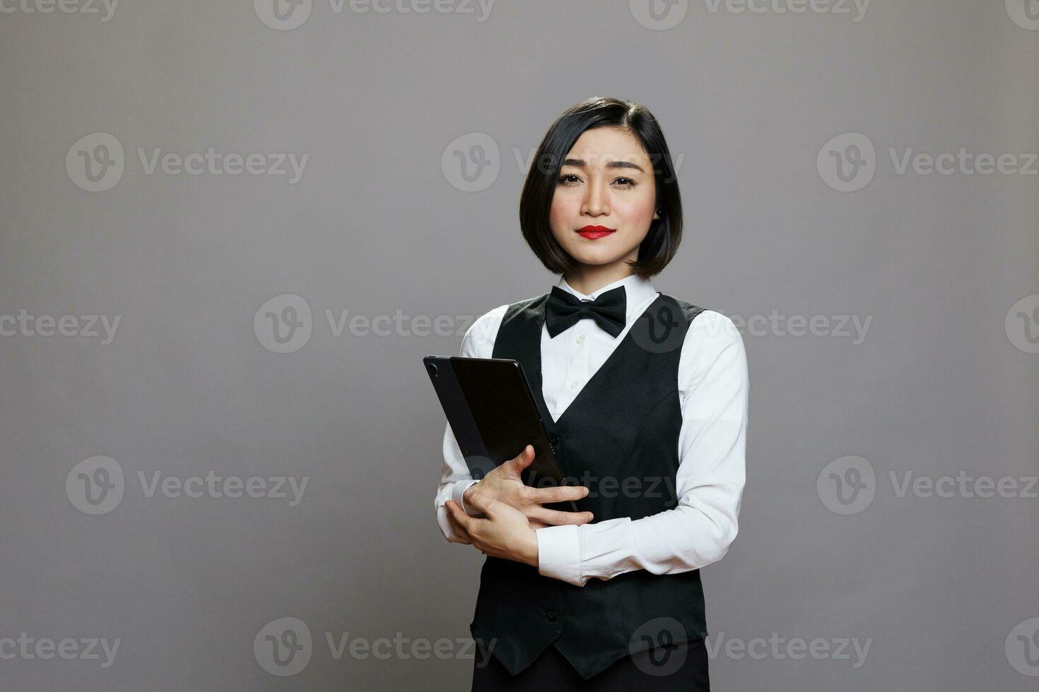 confidente joven asiático mujer camarera vistiendo uniforme y arco Corbata en pie con digital tableta retrato. grave recepcionista participación portátil artilugio en manos y mirando a cámara foto