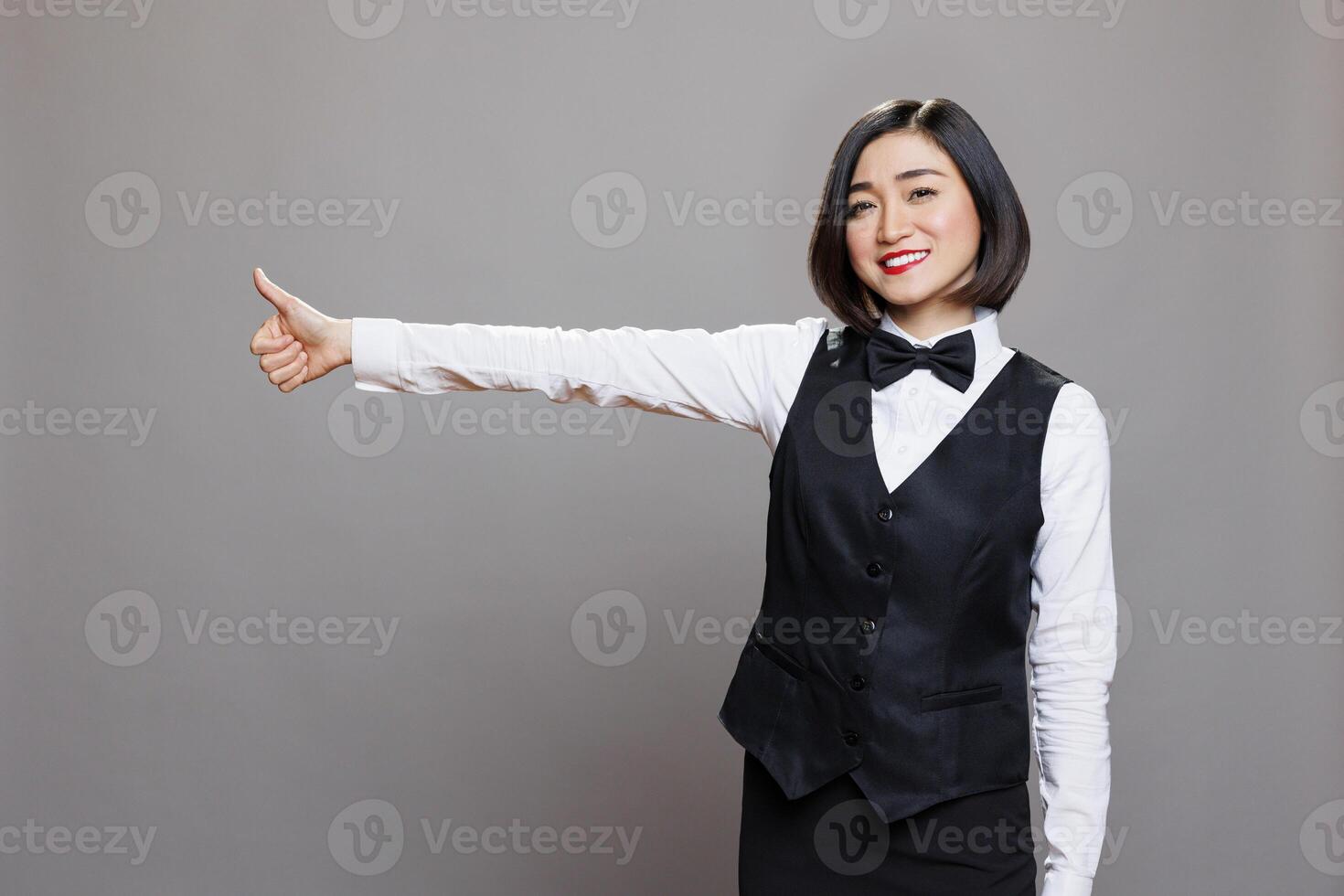 Smiling asian waitress showing thumb up gesture and looking at camera with positive emotion. Cheerful woman receptionist expressing service approval and satisfaction portrait photo