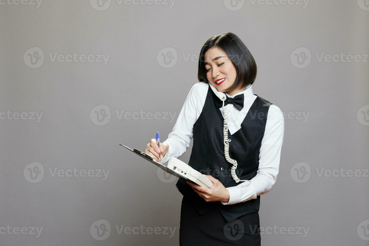 Smiling asian waitress answering landline phone call and writing on clipboard. Restaurant friendly woman employee in uniform talking with client on telephone and managing order photo