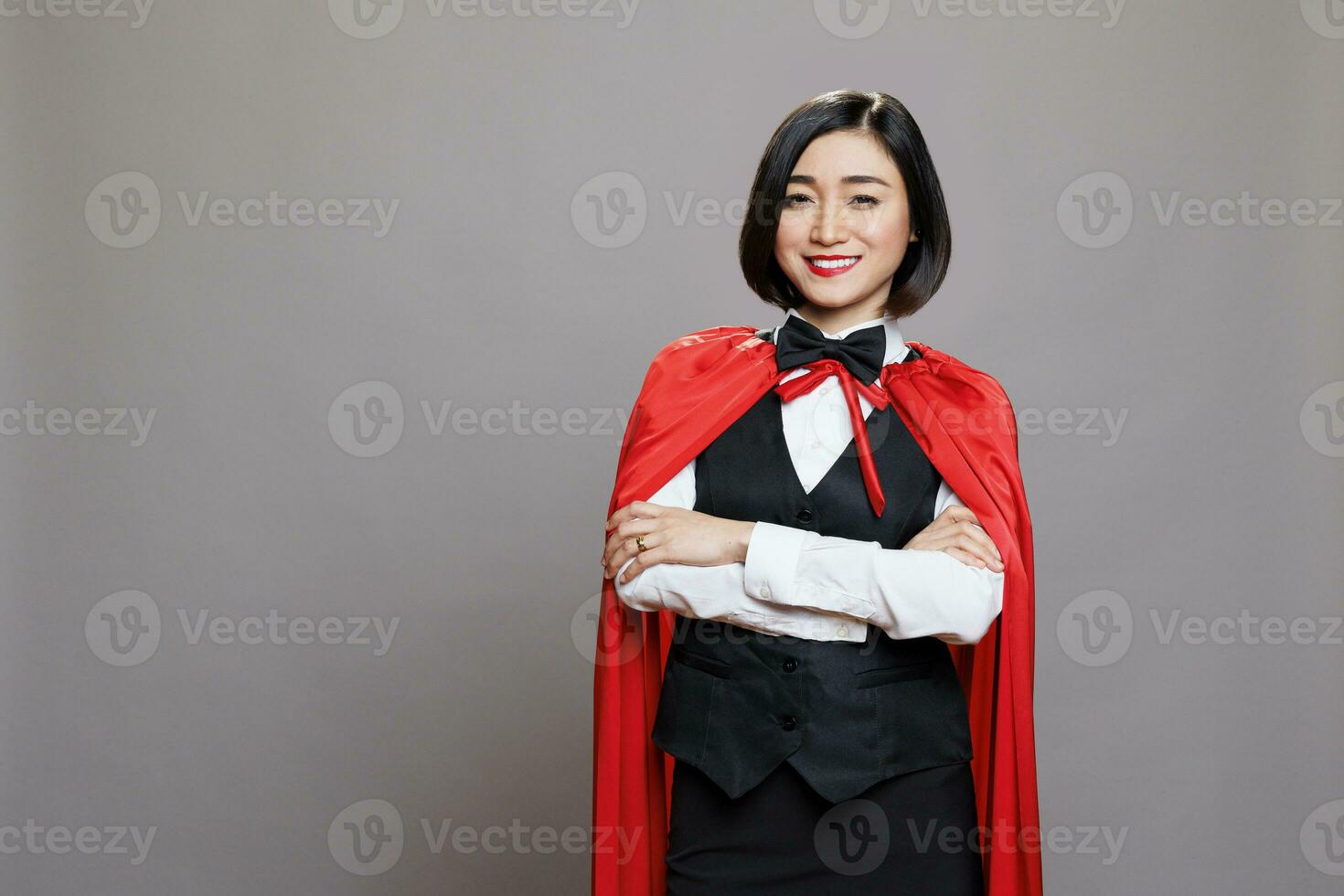 Smiling confident asian waitress in red superwoman cloak standing with arms crossed portrait. Cheerful woman receptionist with folded hands wearing hero cape looking at camera photo