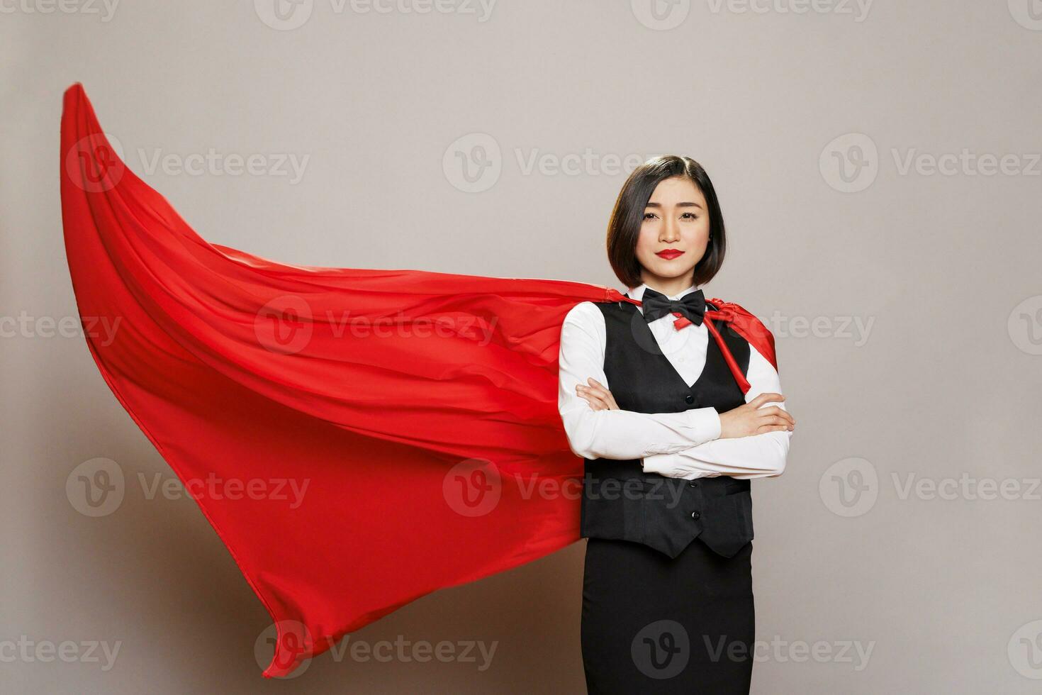 Receptionist with arms crossed wearing red superman cloak, representing power and confidence portrait. Attractive asian waitress dressed in restaurant uniform and fluttering superhero cape photo