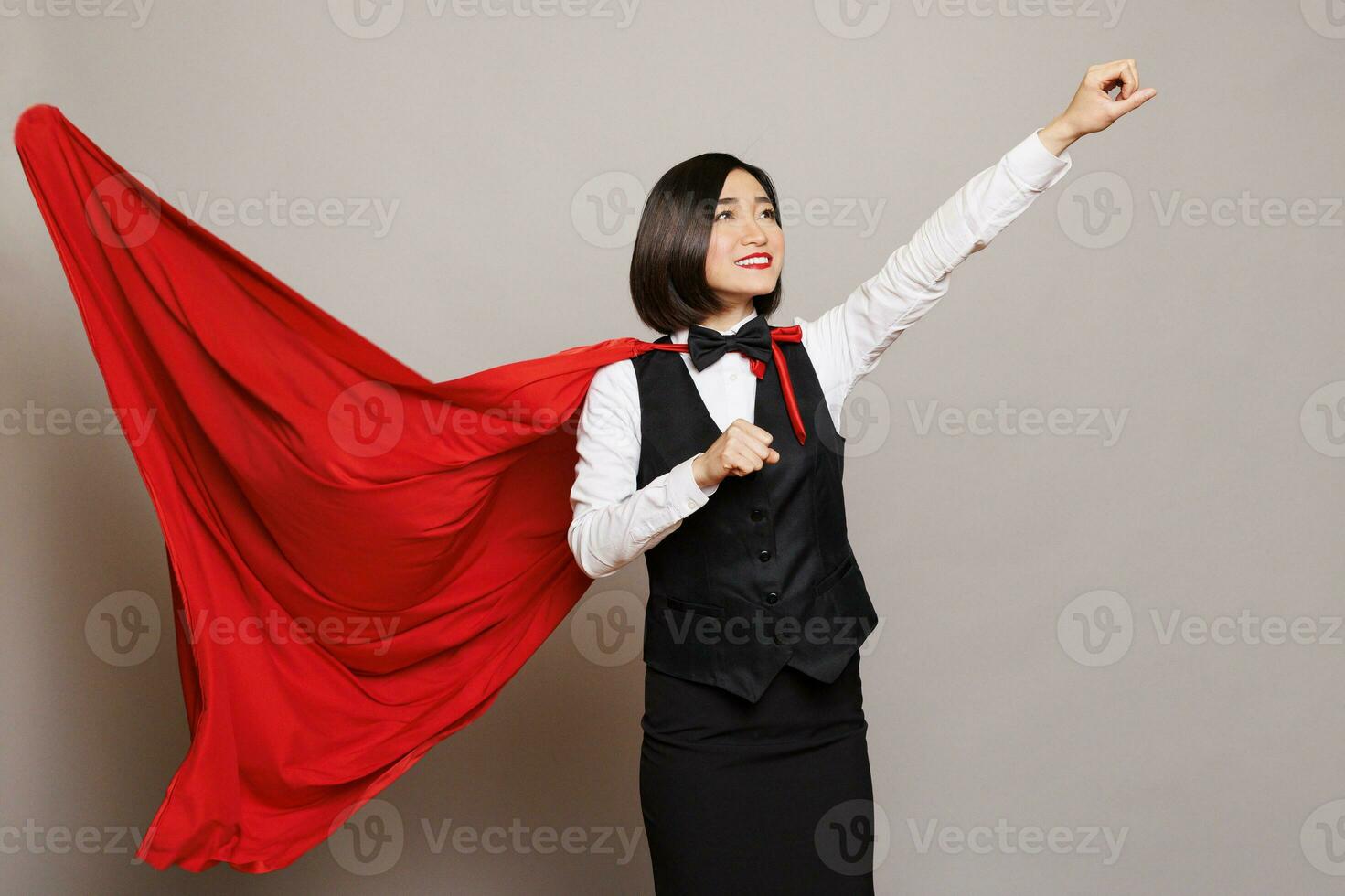 sonriente asiático camarera supermujer en revoloteando capa volador, exhibiendo fuerza y fuerza. alegre confidente recepcionista vistiendo superhombre rojo capa, levantamiento apretado puño y mirando arriba foto