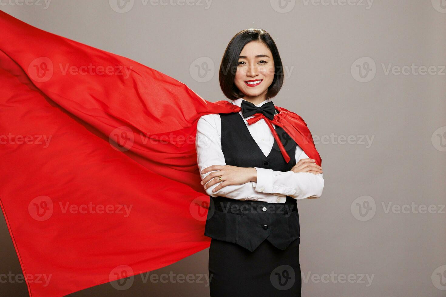 confidente asiático camarera con cruzado brazos en pie en revoloteando rojo héroe capa retrato. sonriente mujer recepcionista vistiendo restaurante uniforme y supermujer capa en estudio foto