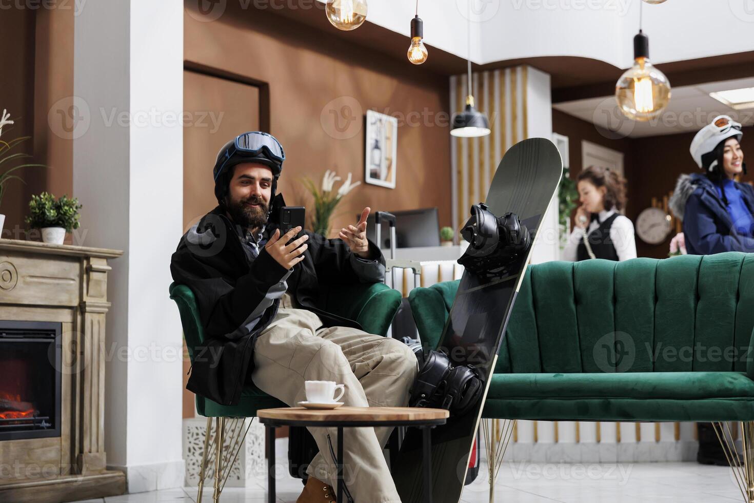 A man seated on sofa browses on his digital phone with snowboard nearby. Caucasian visitor dressed in snow clothing surfing the net using his smartphone. Winter vacation anticipation captured. photo