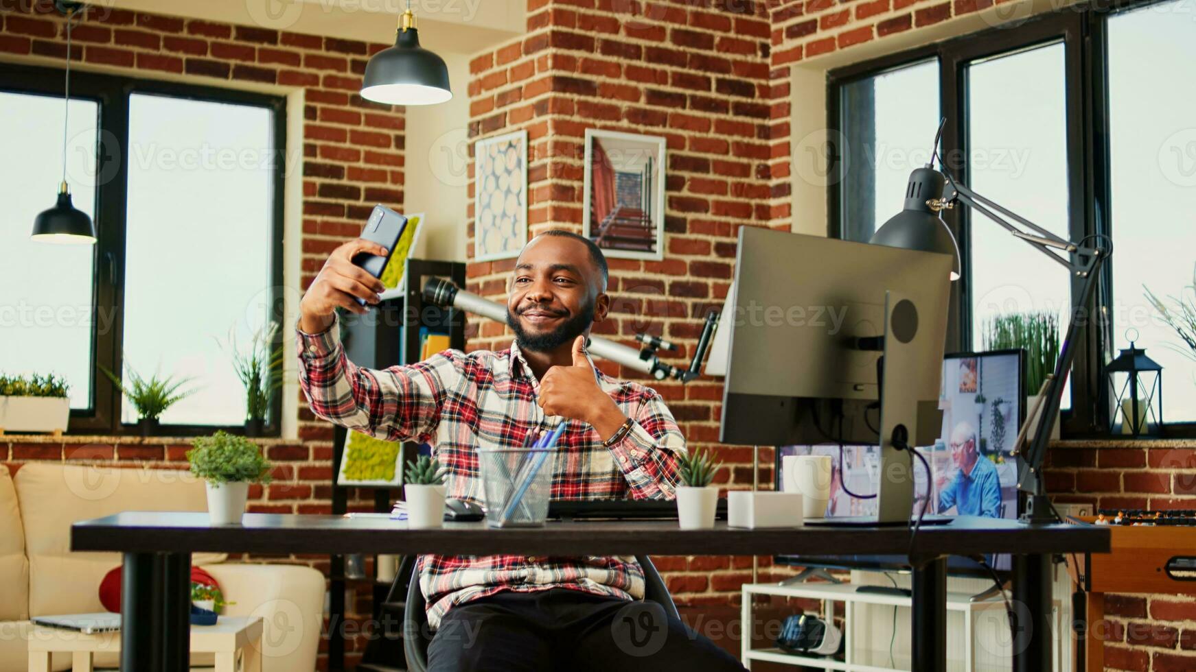 contento alegre africano americano hombre tomando selfie mientras sonriente y demostración pulgares arriba signo. hombre en bonito elegante moderno casa capturar foto utilizando teléfono inteligente mientras remotamente trabajando desde hogar