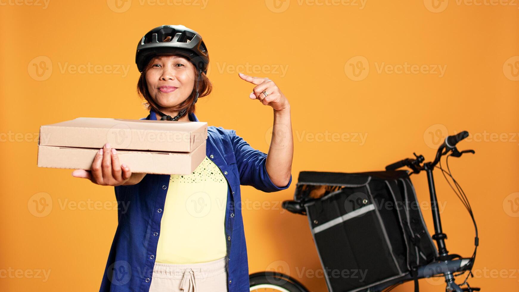 Portrait of joyful professional courier pointing to takeaway pizza boxes. Smiling bike rider showing thumbs up sign while delivering takeout food, isolated over orange studio background photo