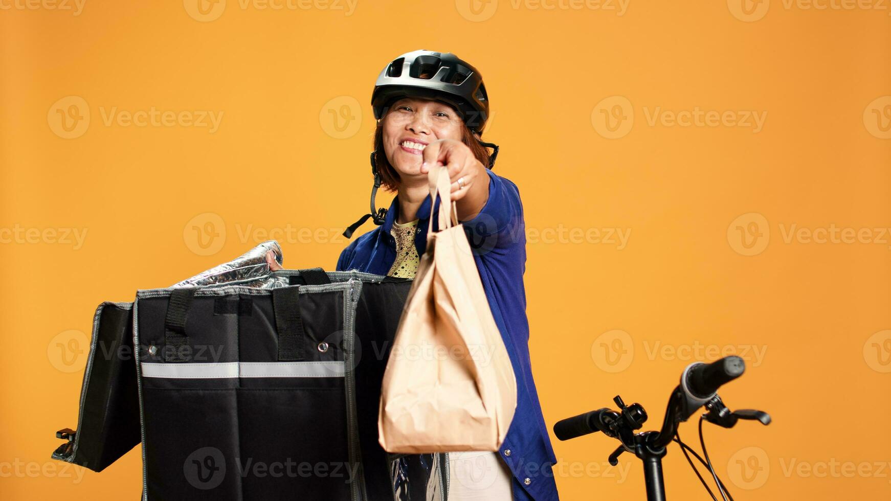 Food delivery bicycle rider preparing meal order while waiting for customer to answer door. Courier isolated over studio background taking lunch bag out of thermal backpack and offering it to client photo
