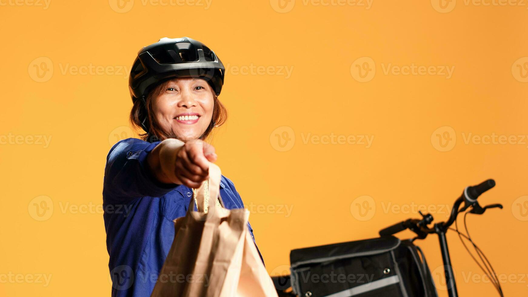 Courier waiting in front of house door for client to pick up takeout meal order, close up. Asian delivery woman offering takeaway food bag to customer, isolated over orange studio background photo